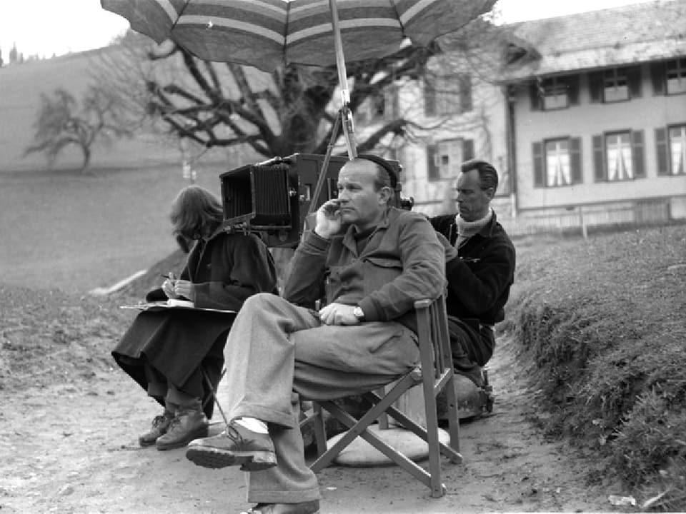 Franz Schnyder sitzt neben der Kamera unter einem Sonnenschrim und betrachtet die Szene. Skript und Kameramann sitzen daneben.