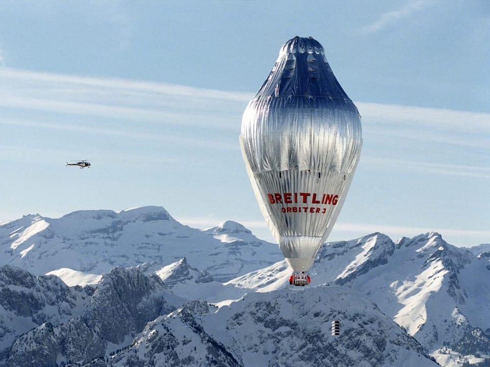 Ein silberner Ballon fliegt vor einem Alpenpanorama. Neben Ballon fliegt ein Helikopter.
