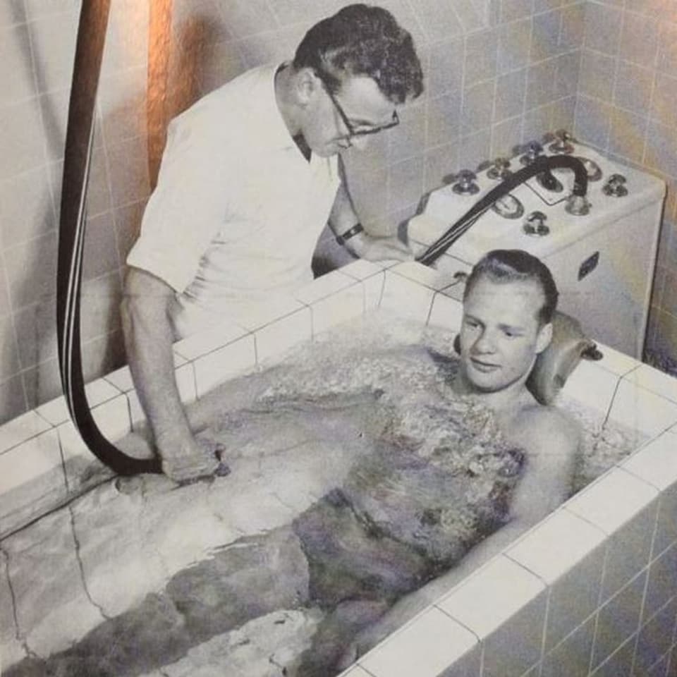 Schwarz-weiss-Aufnahme eines Mannes, der in einer gekachelten Badewanne sitzt.