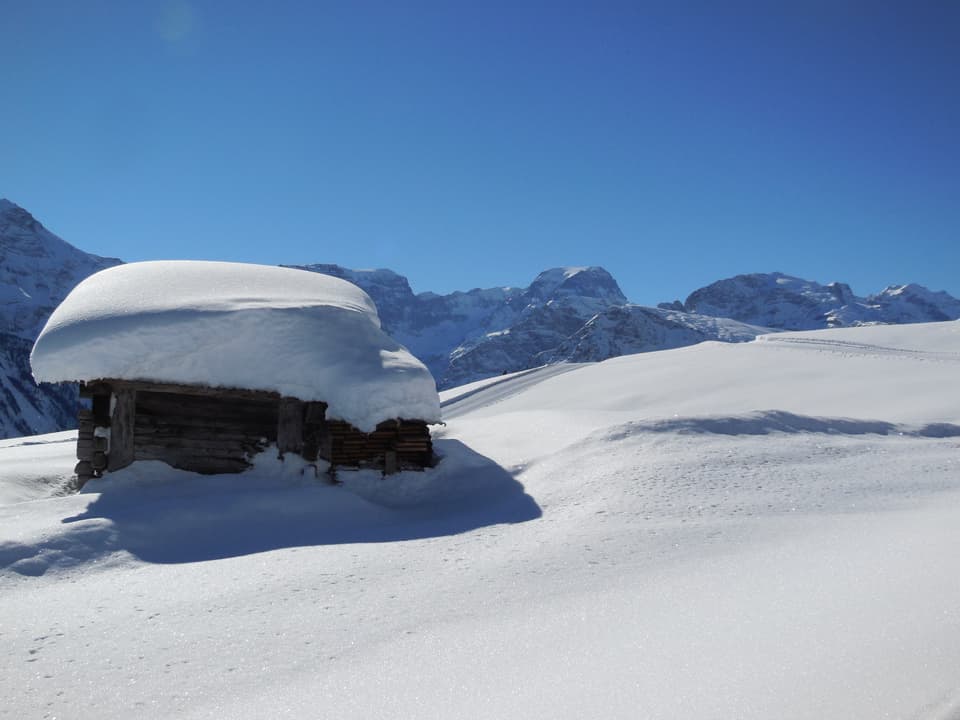 Winterlandschaft: im Vordergrund Holzhütte, im Hintergrund Berge.