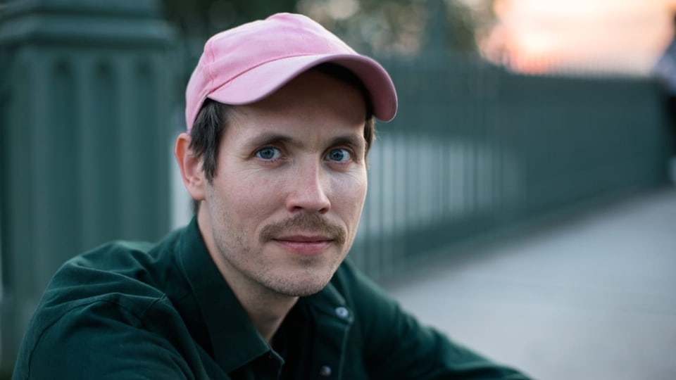 Ein junger Mann mit Schnauz und pinkem Baseball-Cap.