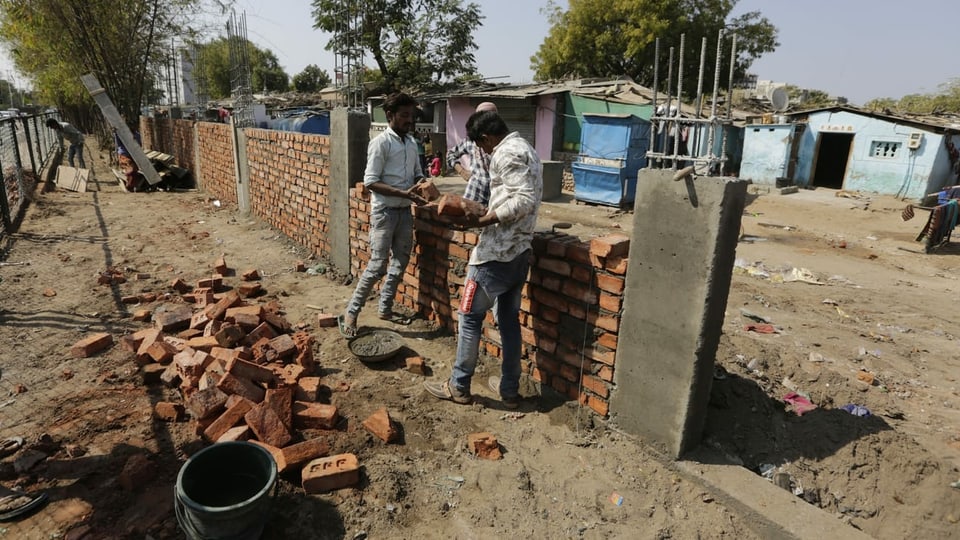Zwei Arbeiter bauen eine Mauer aus Ziegelsteinen. Im Hintergrund ein Slum.