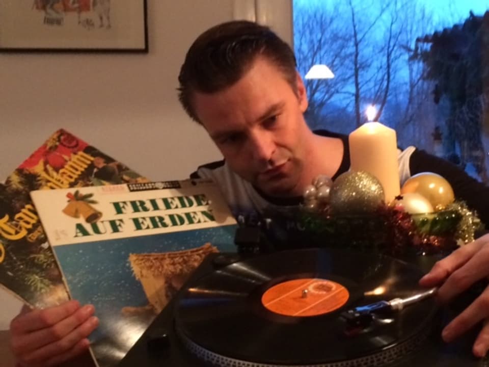 Eric Dauer sitzt vor einer Adventskerze, hält zwei Weihnachtsplatten in der linken Hand, mit der rechten setzt er den Tonabnehmer auf die Schallplatte auf dem Plattenspieler.