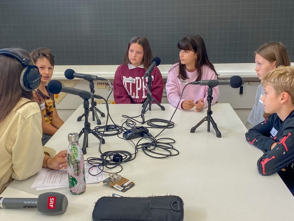 Kinder sitzen rund um einen Tisch und sprechen in Mikrofone