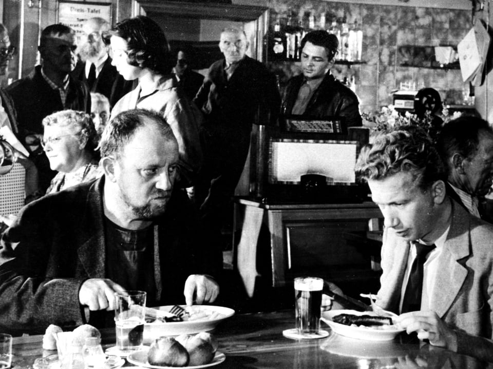 Zwei Männer sitzen an einem Tisch in einer Kneipe und essen. Der Mann mit dem Bart schaut Heinz skeptisch an.