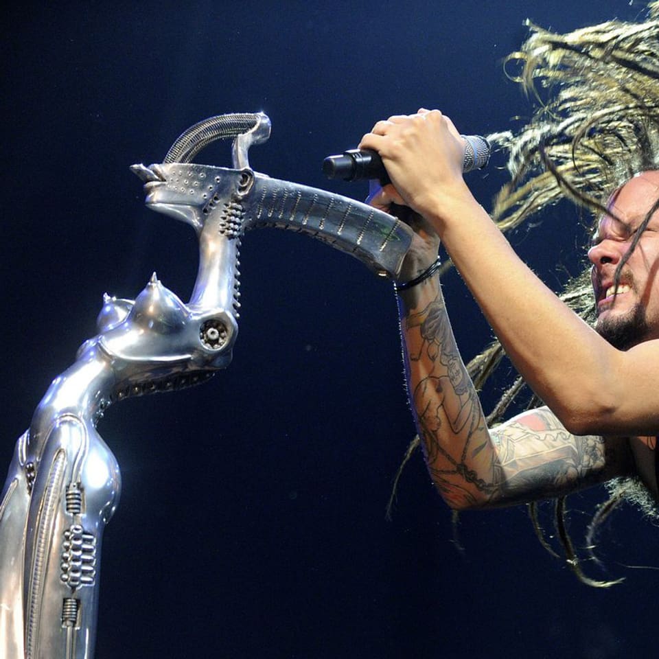 Zu sehen ist Korn-Sänger Jonathan Davis. Er ist am Headbangen. Vor ihm steht ein alieneske Frauengestalt aus Metall. Es ist der Mikrofonständer.