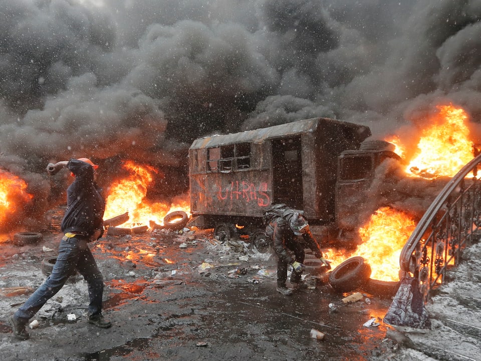 Feuer, Rauch, ausgebrannter Lastwagen, davor ein Demonstrant, der einen Stein wirft.
