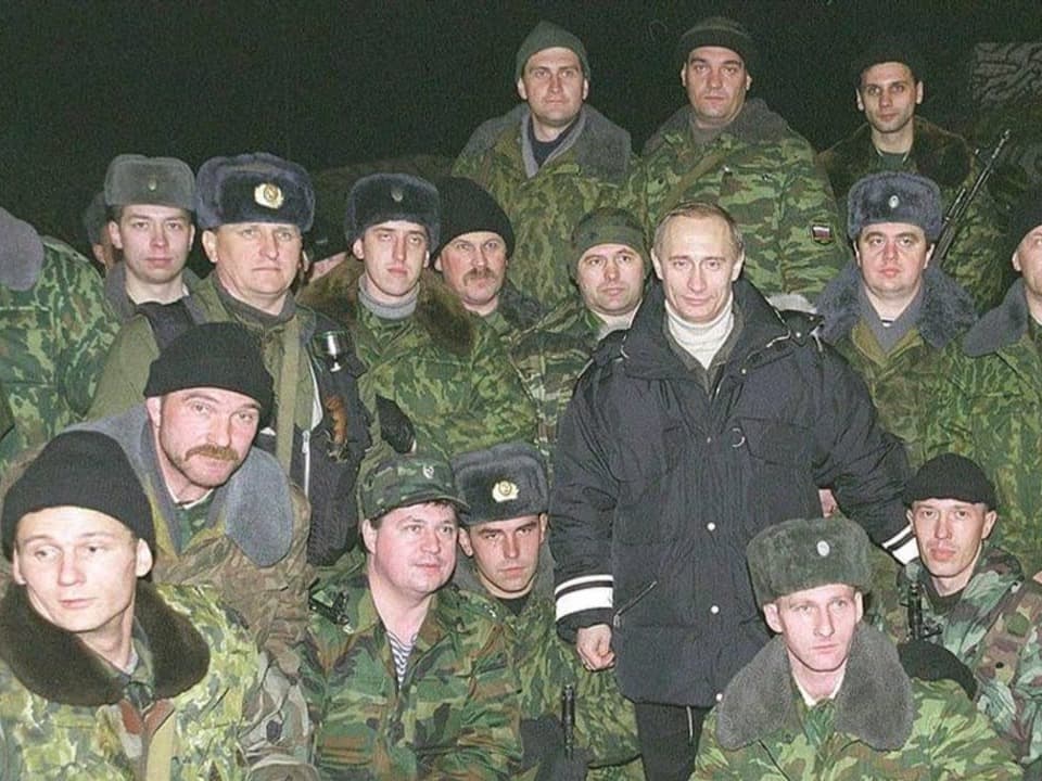 Ein Mann in schwarzer Jacke (Putin) steht inmitten von Soldaten in Camouflage-Kleidung