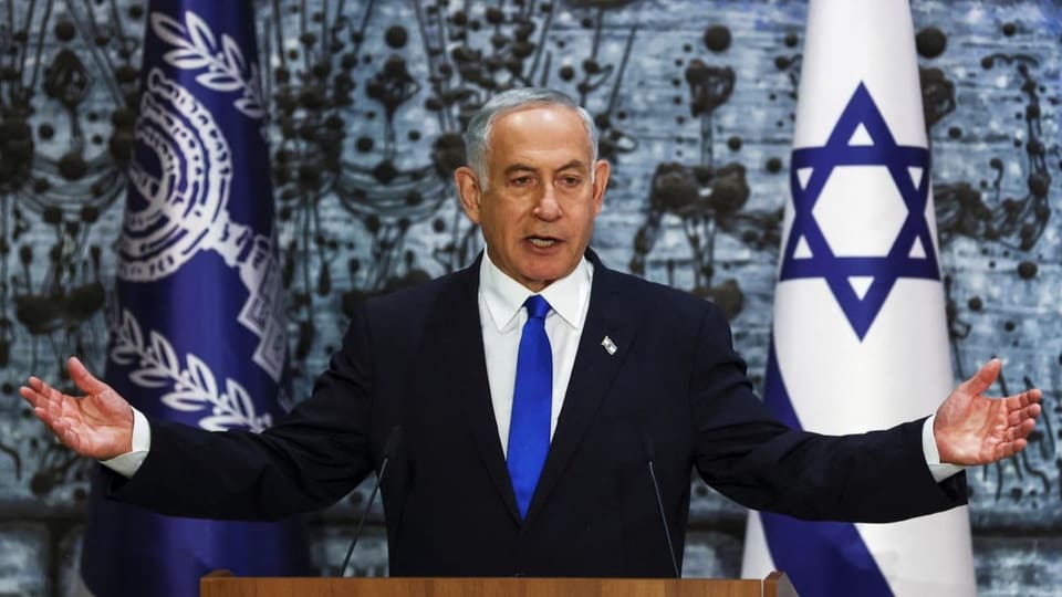 Auf dem Bild steht Benjamin Netanjahu mit ausgebreiteten Händen am Rednerpult.