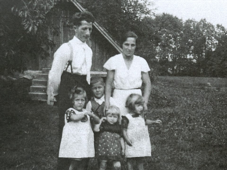 Schwarz-Weiss-Fotografie mit einer sechsköpfigen Familie, die auf einer Wiese vor einem Holzhaus steht.