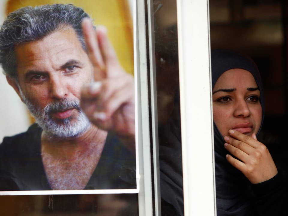 Auf dem Poster ist Mer Khamis zu sehen, wie er das Victory-Zeichen macht. Die Frau neben dem Poster ist schwarz gekleidet und blickt traurig.