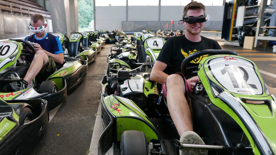 Zwei FPV-Racer sitzen in Go-Karts, mit Display-Brillen, und steuern ihre Quadrokopter.