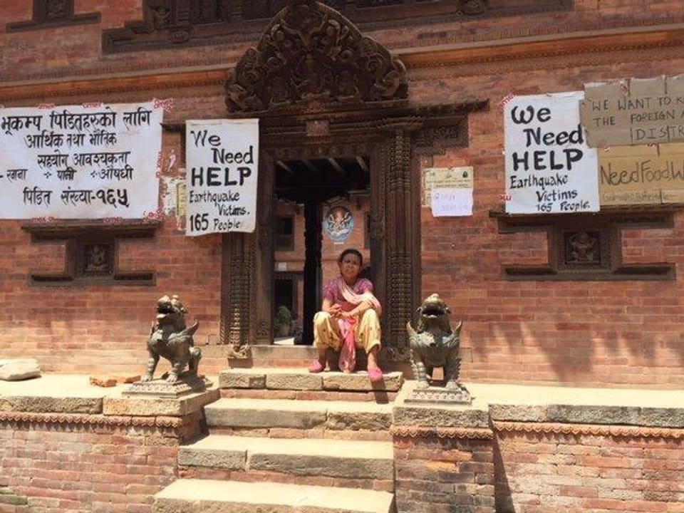 Eine Frau sitzt vor einem Gebäude. Auf Plakaten steht auf Englisch «wir brauchen Hilfe».
