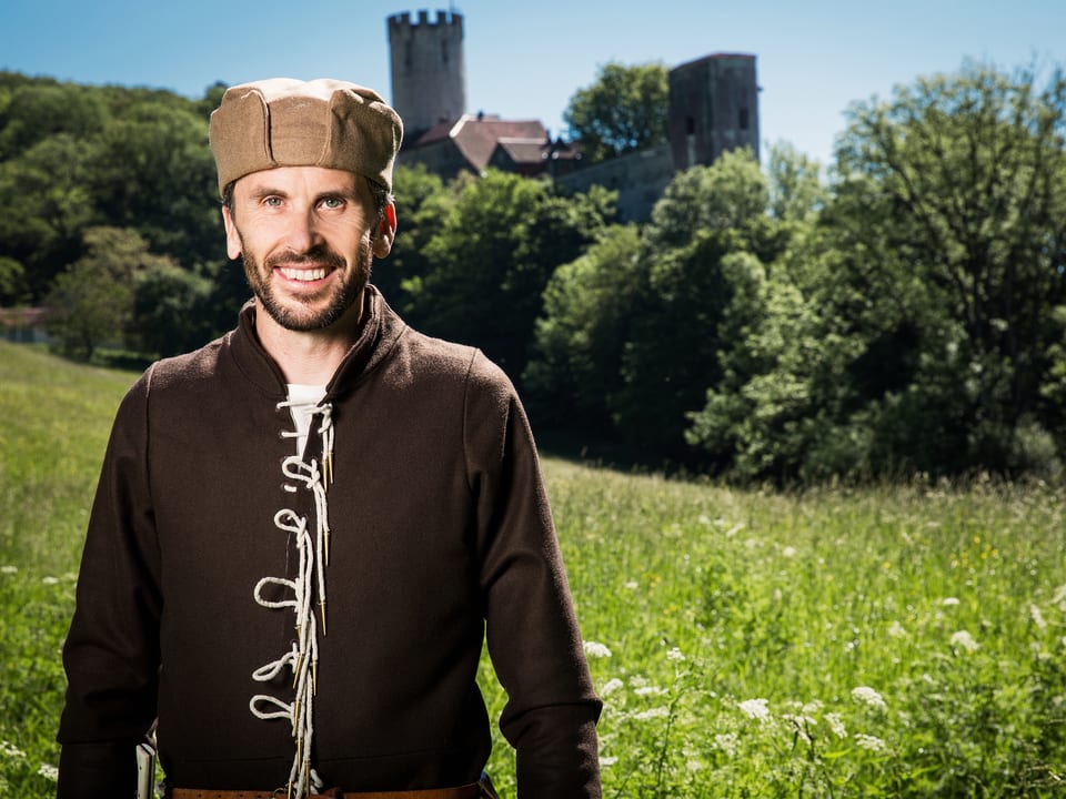 Mann in mittelalterlicher Kleidung auf einer Wiese mit einer Burg im Hintergrund