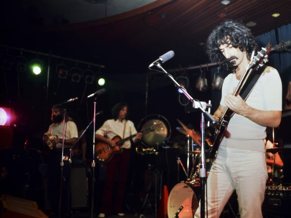 Mann mit langen Haaren und Ziegenbart (Frank Zappa) spielt Gitarre, im Hintergrund zwei Bandmitglieder