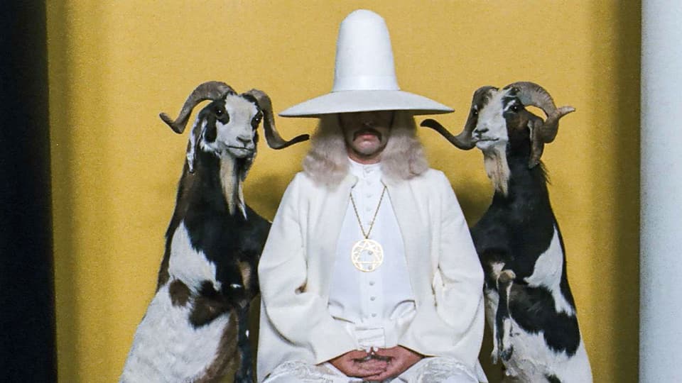 Mann in Robe. Ein Hut verdeckt die Hälft seines Gesichts. Links und rechts stehen Ziegen.