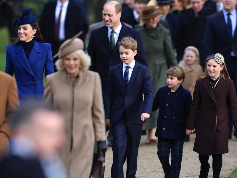 Man sieht Kate und William und ihre drei Kinder. Sie sind am laufen. Vor ihnen is Königin Camilla.