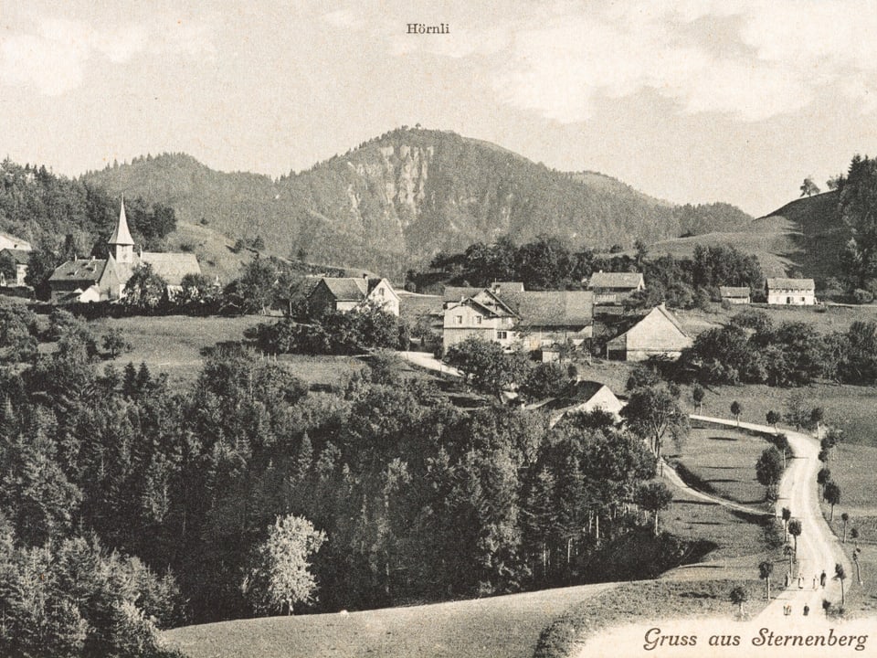 Eine Postkarte in schwarz-weiss aus Sternenberg. Im Vordergrund eine staubige Strasse, hinten ein paar Häuser und das Kirchlein.