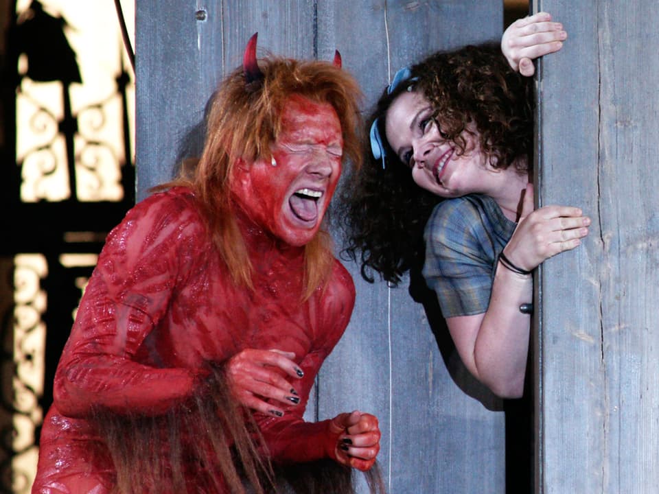 Eine rote Teufelfigur steht mit geschlossenen Augen und ausgestreckter Zunge neben einer Jungen Frau, die lächelnd hinter einer Bretterwand hervorsdchaut.