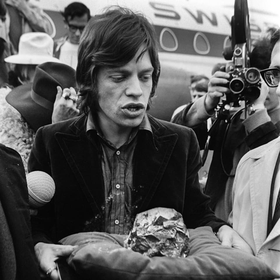 Mick Jagger 1967 am Flughafen Zürich. Er hält ein Kissen mit einem Stein darauf vor sich.