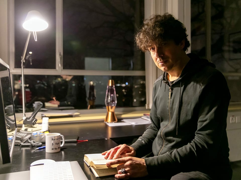 Ein Mann in einem dunklen Büro, das nur von einer Tischlampe beleuchtet wird. Er sitzt an einem Tisch und blättert in einem Buch.