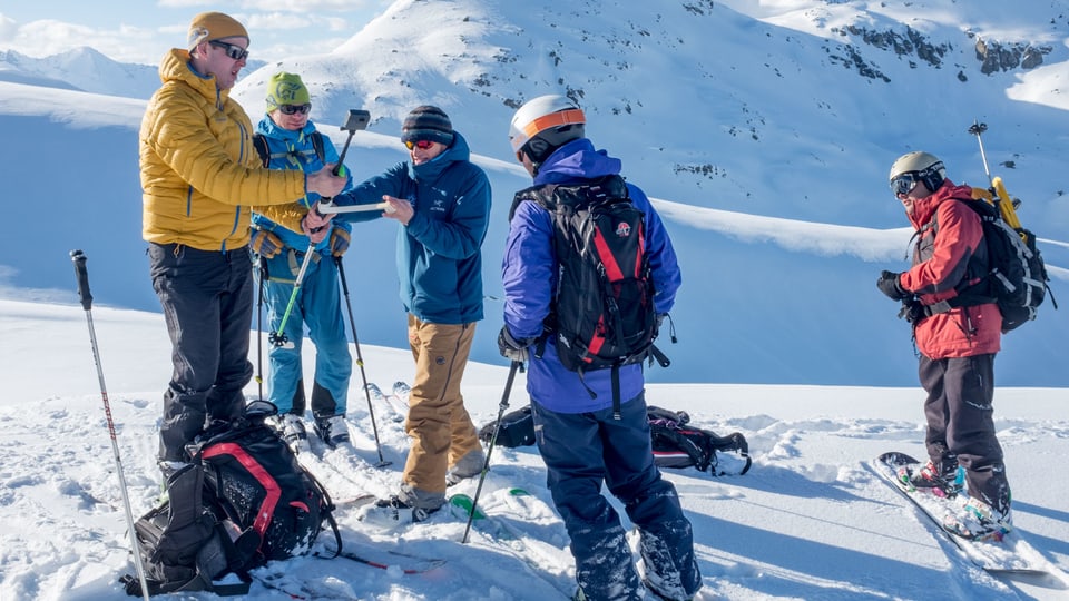 Eine Gruppe von Wintersportlern steht in einer verschneiten Berglandschaft. Zwei Menschen befestigen eine Kamera mit Klebeband and einem Skistock.