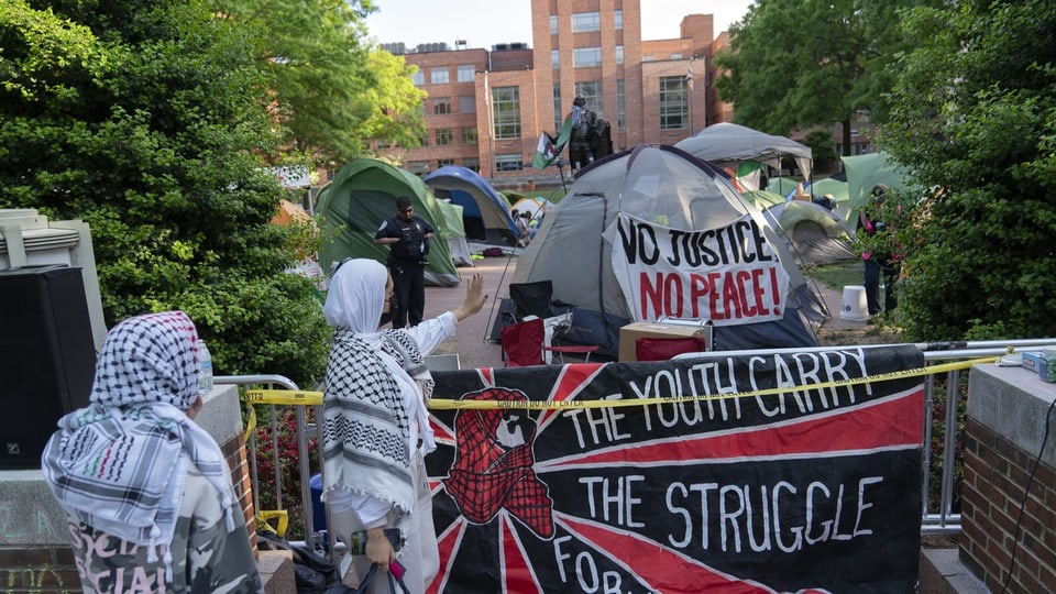 Protestcamp mit Zelten und Bannern, darunter 'No Justice No Peace', in städtischem Park.