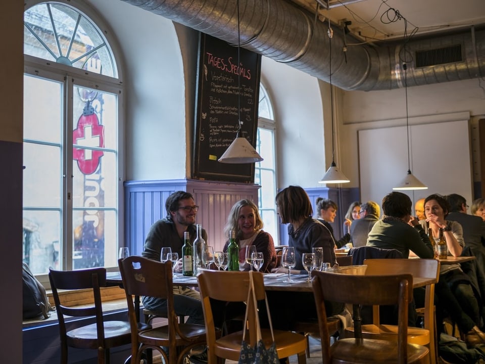Menschen beim Essen in einem belebten Restaurant mit grosser Fensterfront und Tafel mit Tages-Specials.