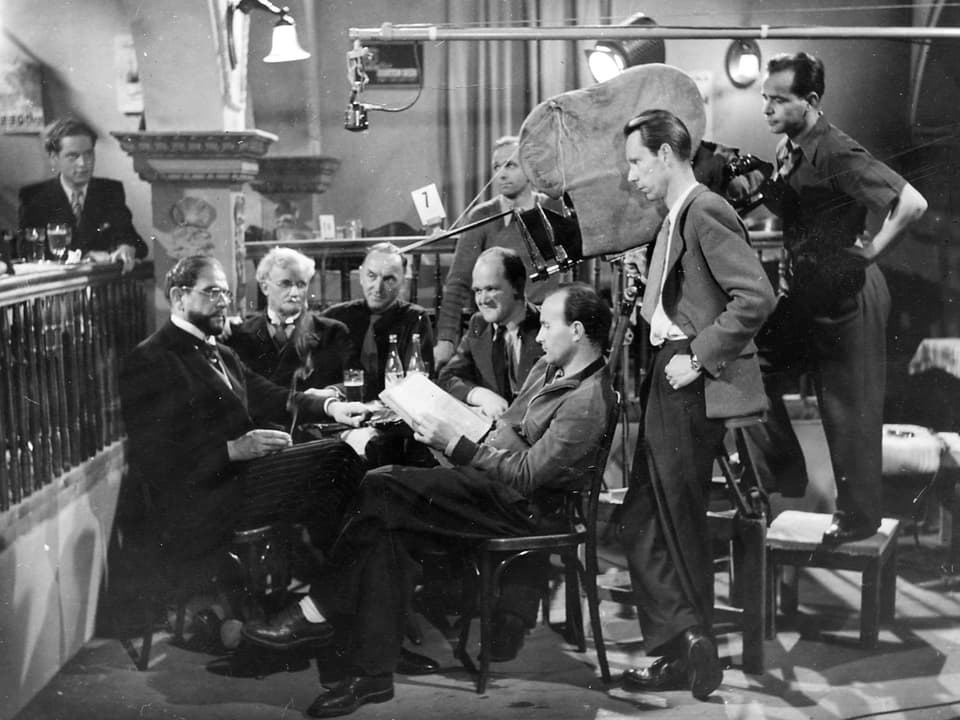 Um einen Tisch in einem Restaurant sitzen fünf Schauspieler. Daneben steht die Kamera mit dem Kameramann, dem Regisseur und dem Tonmann.