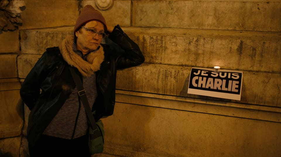 Frau an Wand lehnend, Daneben ein Plakat darauf steht: «Je suis Charlie»