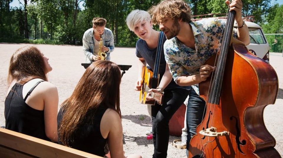 Zwei Frauen sitzen auf einer Parkbank und werden von drei Musikern mit Gitarre, Kontrabass und Saxophon mit einem Ständchen überrascht.