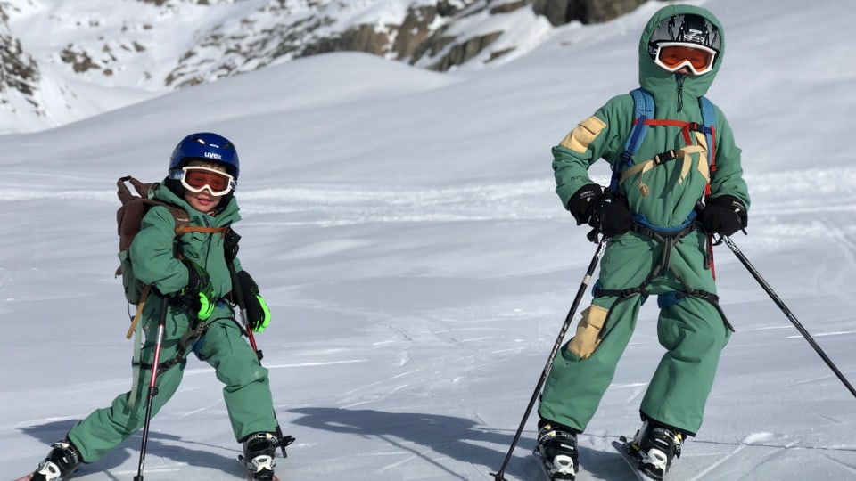 Zwei Jungen sind mit Tourenskiern unterwegs. Sie tragen beide einen grünen Schneenazug und stehen im Schnee. 