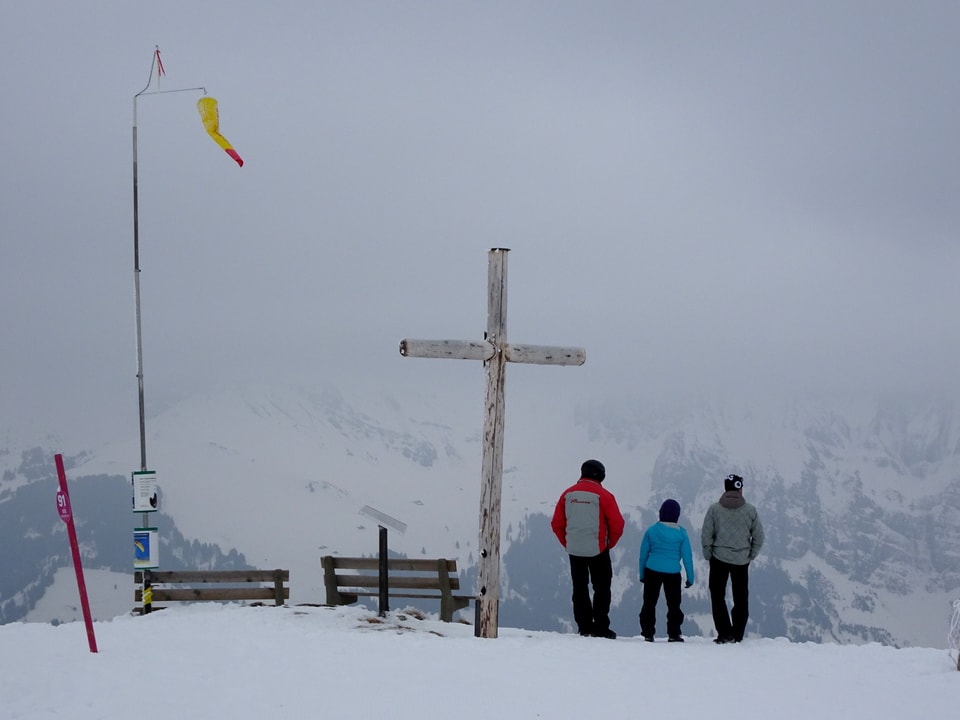 Grauer Himmel, ein Bergkreuz, 2 Bänke, 3 Personen mit Rücken zum Fotografen