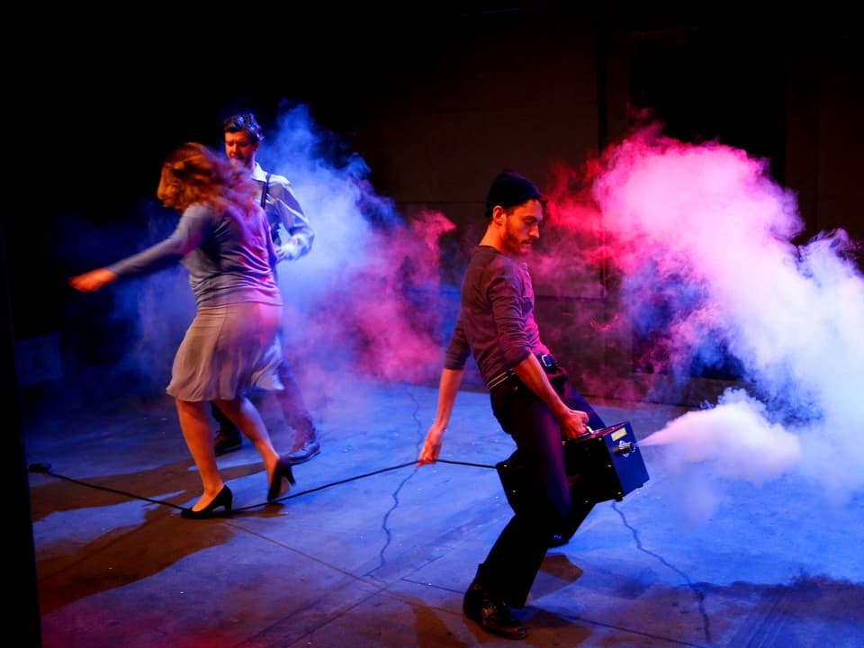 Eine Frau und zwei Männer auf der Bühne, Rauch und lila Licht, einer der Männer reitet auf einem Transistor.