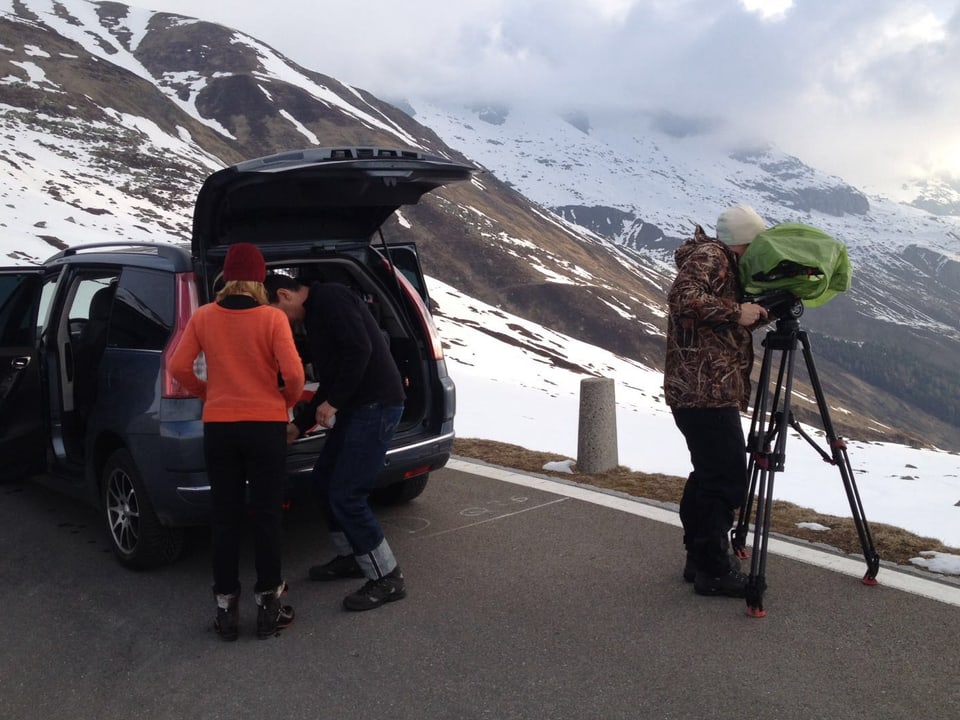 Ein offenes Auto, daneben steht ein Kameramann und filmt die Landschaft. Ein Mann nimmt etwas aus dem Kofferraum, daneben wartet eine Frau.