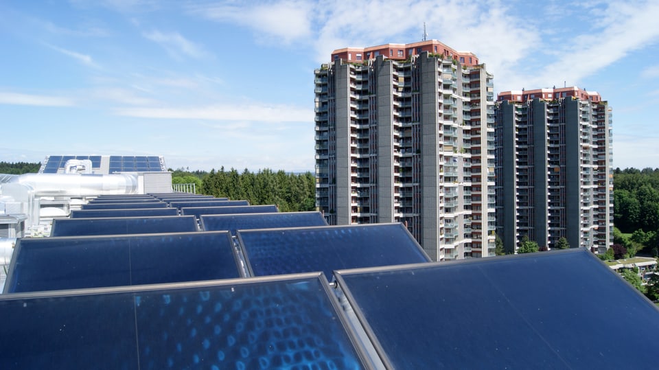 Solarzellen auf einem Dach. Im Hintergrund Hochhäuser.