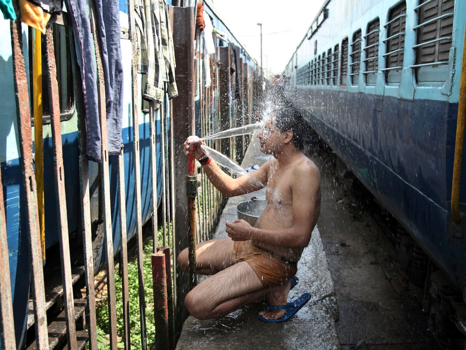 Ein Mann sitzt auf einem Betonweg zwischen zwei Zügen und spritzt sich Wasser ins Gesicht.