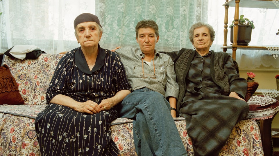 Eine Frau mit kurzen Haaren und männlicher Kleidung sitzt zwischen zwei älteren Frauen auf einem Sofa.