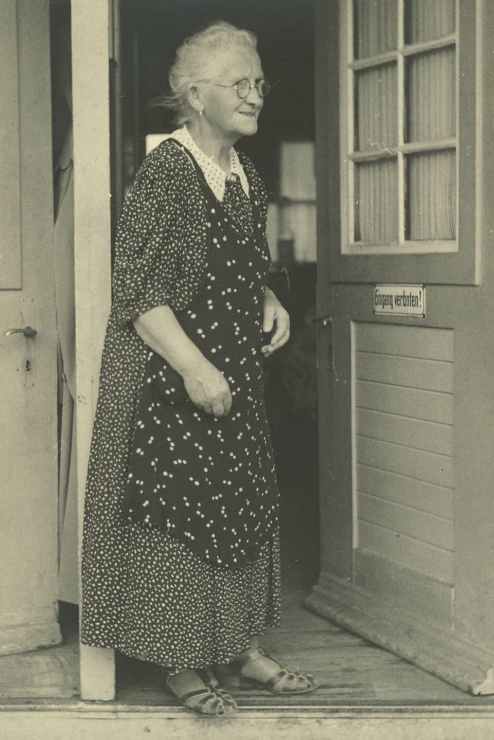 Schwarz-Weiss-Fotografie von einer alten Frau mit Brille, die vor einer offenen Türe steht.