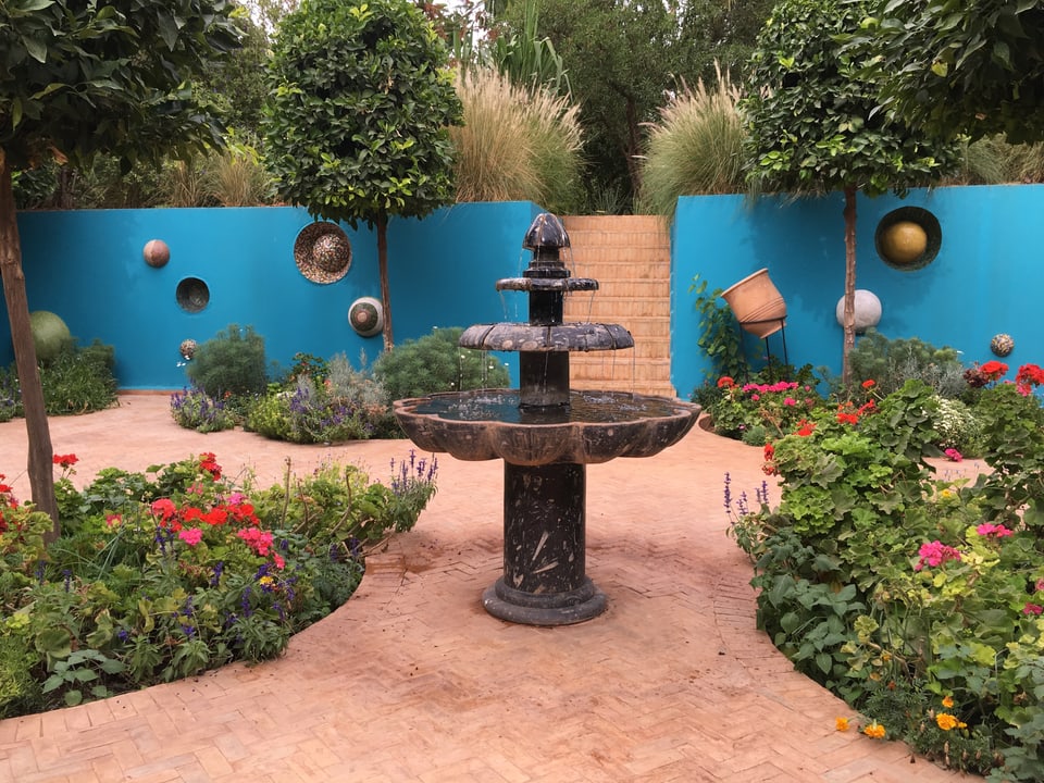 Ein Brunnen vor einer türkisen Wand, der umhüllt ist von Planzen und Blumen. 