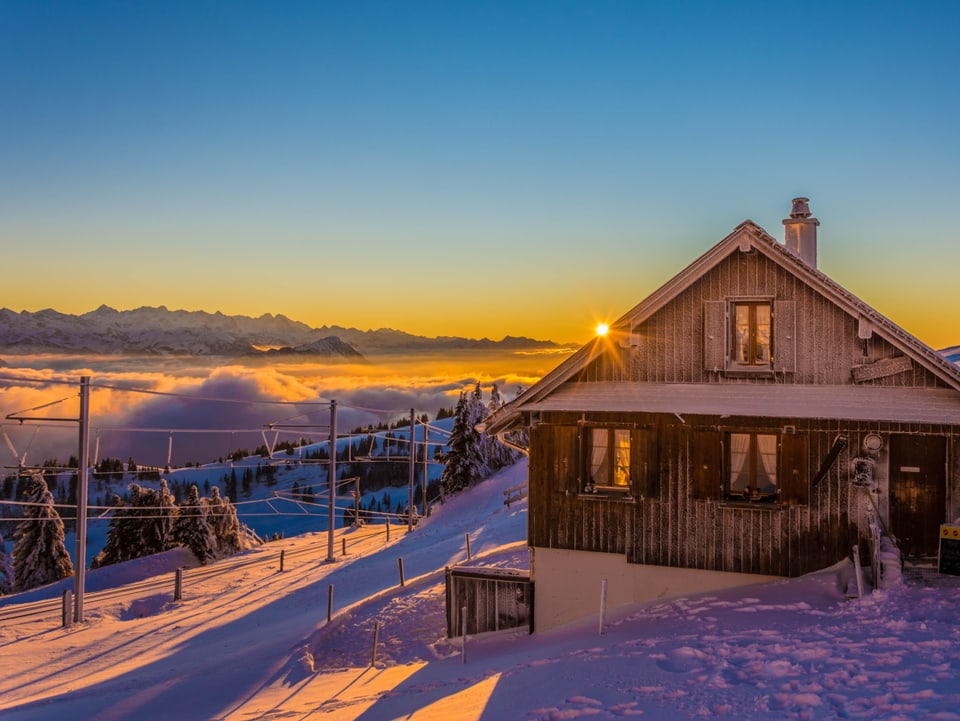Haus auf einem tief verschneiten Berg. Die Sonne geht unter und der Himmel ist oben wolkenlos, während unten im Tal Hochnebel liegt. 