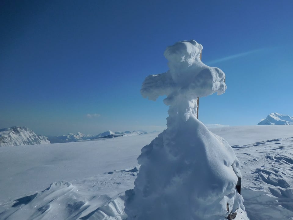 Das Gipfelkreuz ist nicht zu erkennen, vorne ist es mit Schnee bedeckt. Dahinter sind Schneeberge und blauer Himmel zu sehen.