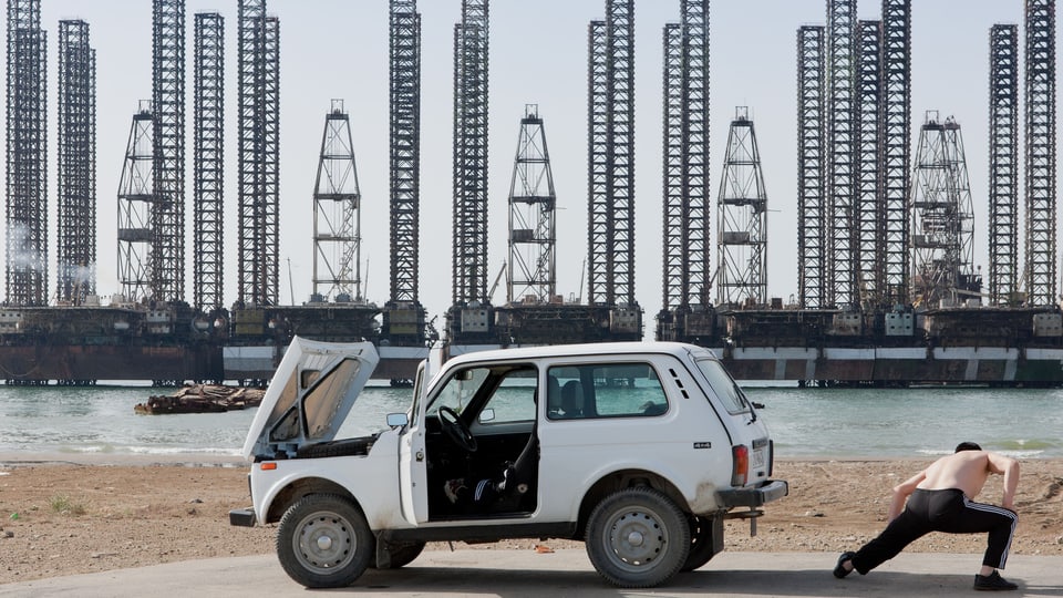 Ein Mensch mit nacktem Oberkörper dehnt sich neben einem Lada-Jeep. Im Hintergrund stehen skelettartige Stahltürme.