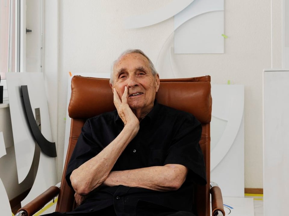 Gottfried Honegger, aufgenommen an seinem 95. Geburtstag, sitzt in einem Sessel in seinem Atelier, Werke von ihm im Hintergrund an der Wand.