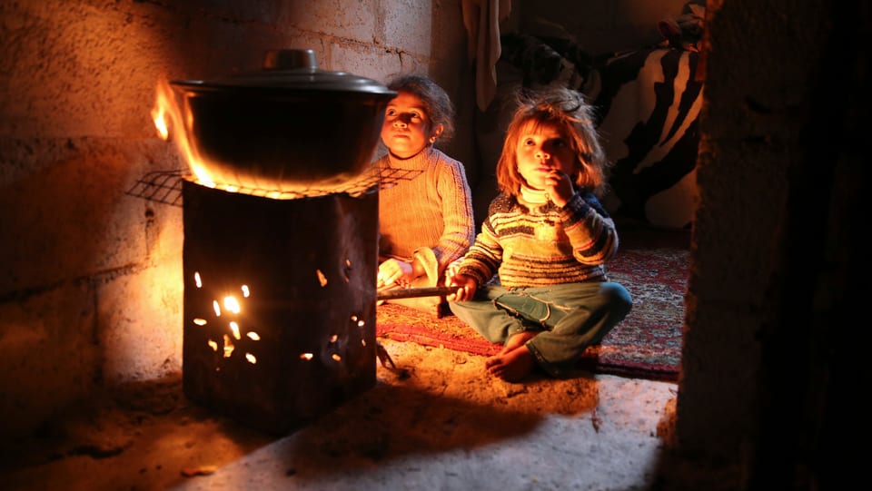 Kinder sitzen im in einem Zimmer um einen Ofen mit offenem Feuer.