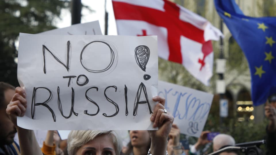 Person hält Plakat mit der Aufschrift «No to Russia» hoch; dahinter georgische und EU-Flagge