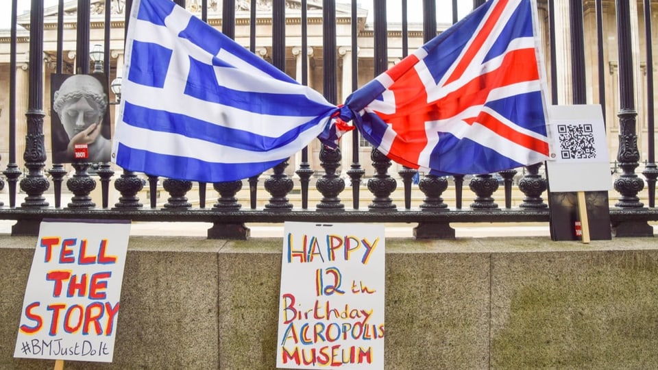 Eine griechische und eine UK-Flagge hängen aneinander geknüpft  an einem Zaun vor einem grossen Museumsgebäude.