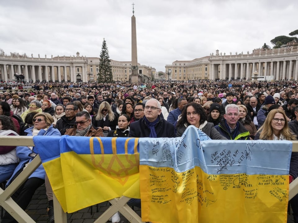 Meschen im Vatikan lauschen der Rede des Papstes vor zwei ukrainischen Flaggen.