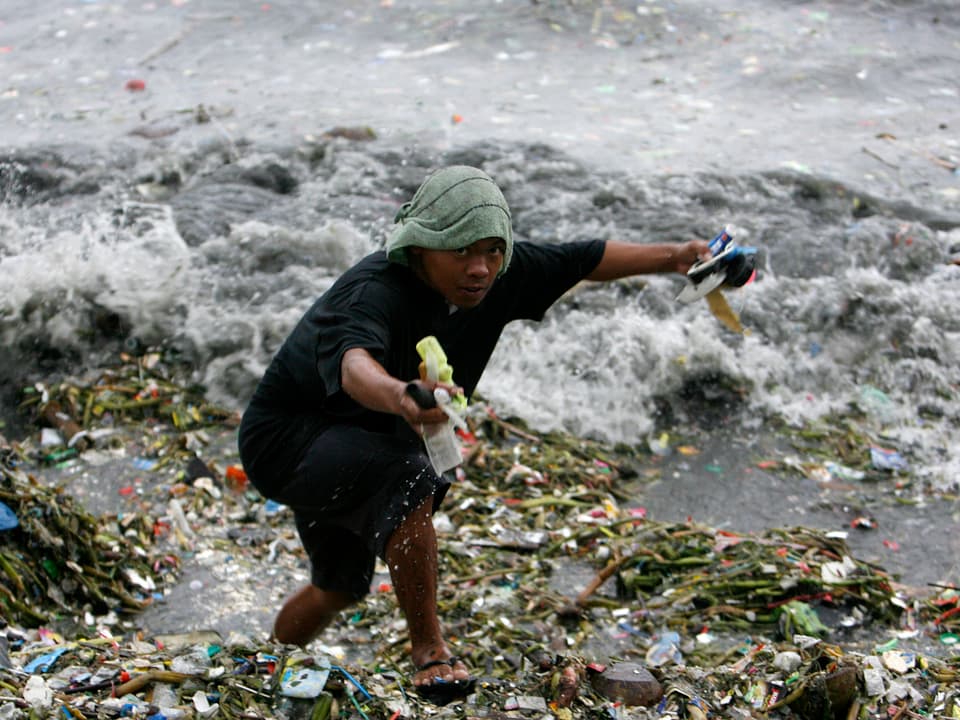 Ein Philippino klettert am Strand über einen Müllberg und hält Plastikabfälle in den Händen.