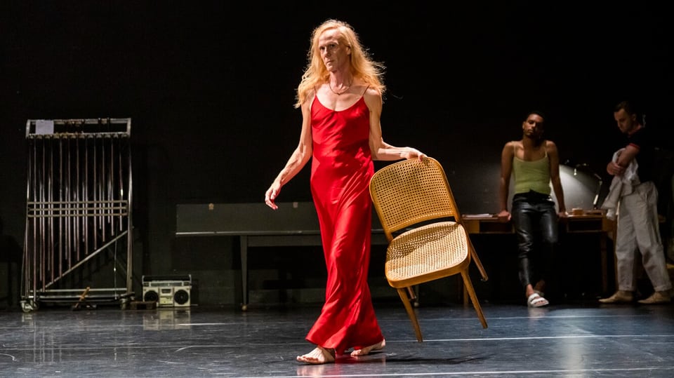 Eine schlanke Person mit langem Haar und rotem langen Kleid steht auf einer Bühne und hält einen Stuhl in der Hand.